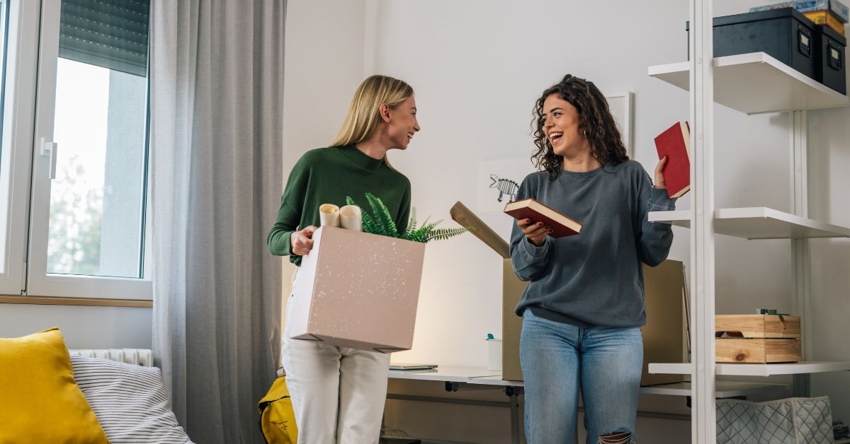 Two young ladies talking one holding a book another with box in a collge dorm